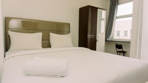 A neatly made bed with white linens and a folded towel, a dark wooden wardrobe, and a chair near a window with a view of buildings. - Studio With Comfortable Design At Serpong Garden Apartment (Cisauk)