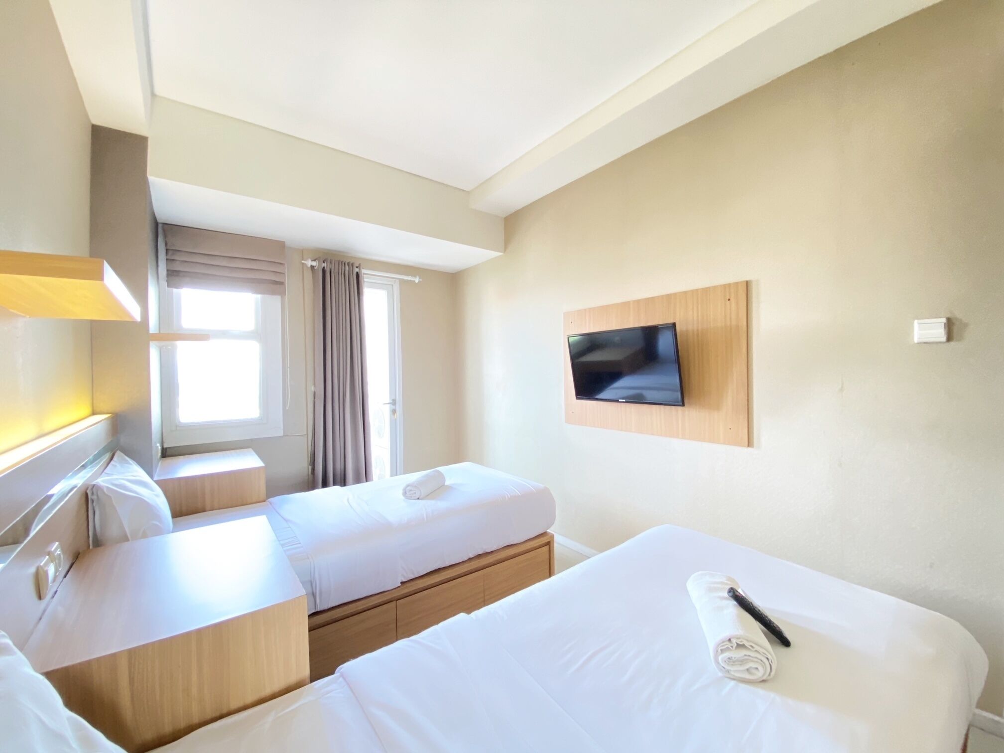 Ein Hotelzimmer mit zwei Betten, einem an der Wand montierten Fernseher, einem Fenster mit Vorhängen und einem Nachttisch.
