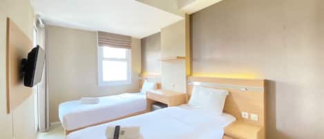 Ein Hotelzimmer mit zwei Einzelbetten, einem an der Wand befestigten Fernseher, einem Nachttisch mit Lampe und einem Fenster mit Vorhängen.
