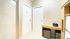 Une petite pièce propre, avec des murs blancs et un sol carrelé. On y trouve deux portes, une petite table en bois avec un distributeur d’eau et un réfrigérateur.