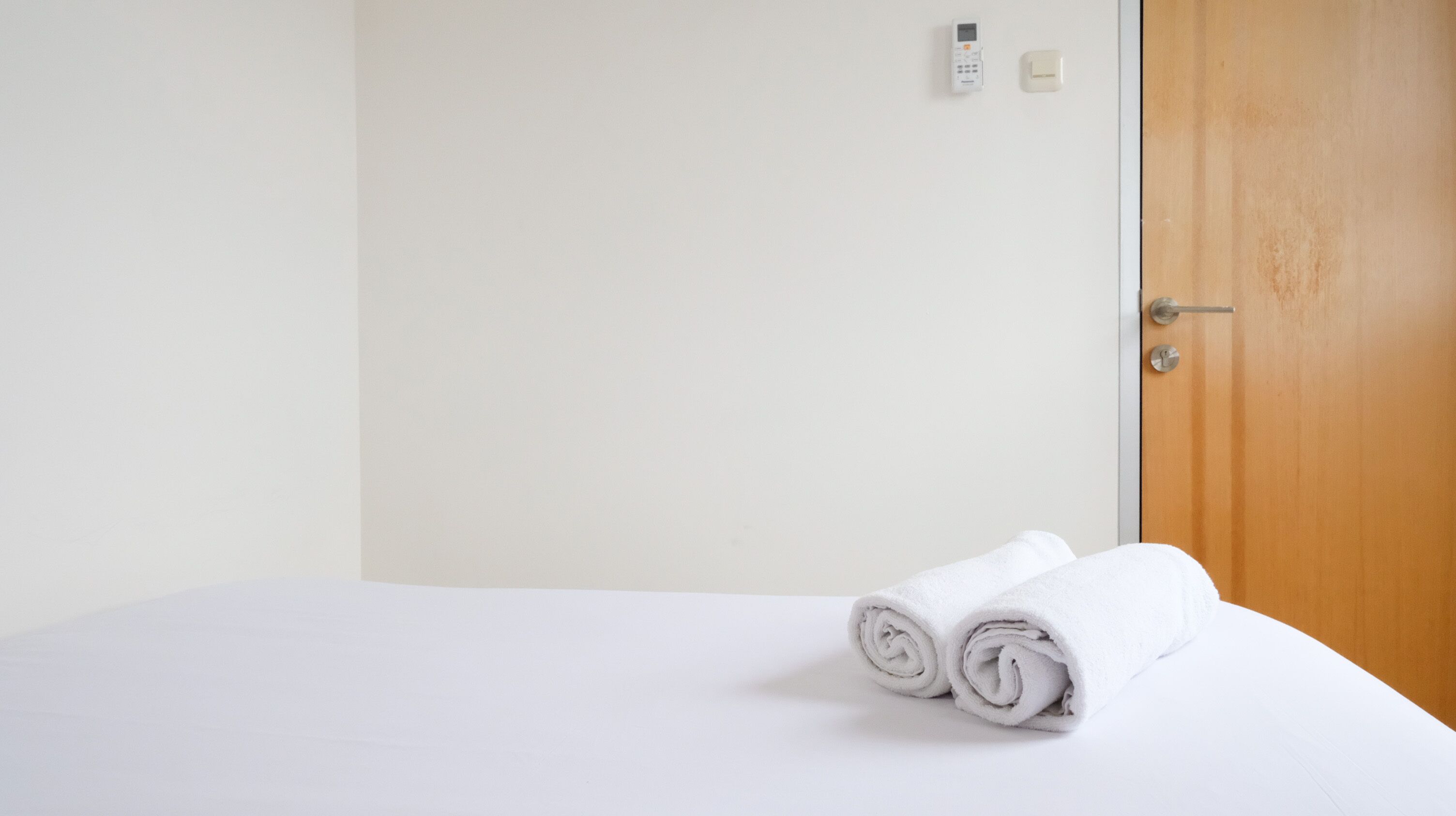 Un letto rifatto con asciugamani bianchi arrotolati, una porta in legno e un termostato fissato al muro.