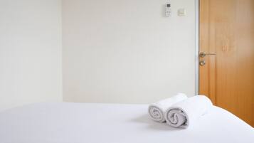 Un letto rifatto con asciugamani bianchi arrotolati, una porta in legno e un termostato fissato al muro.