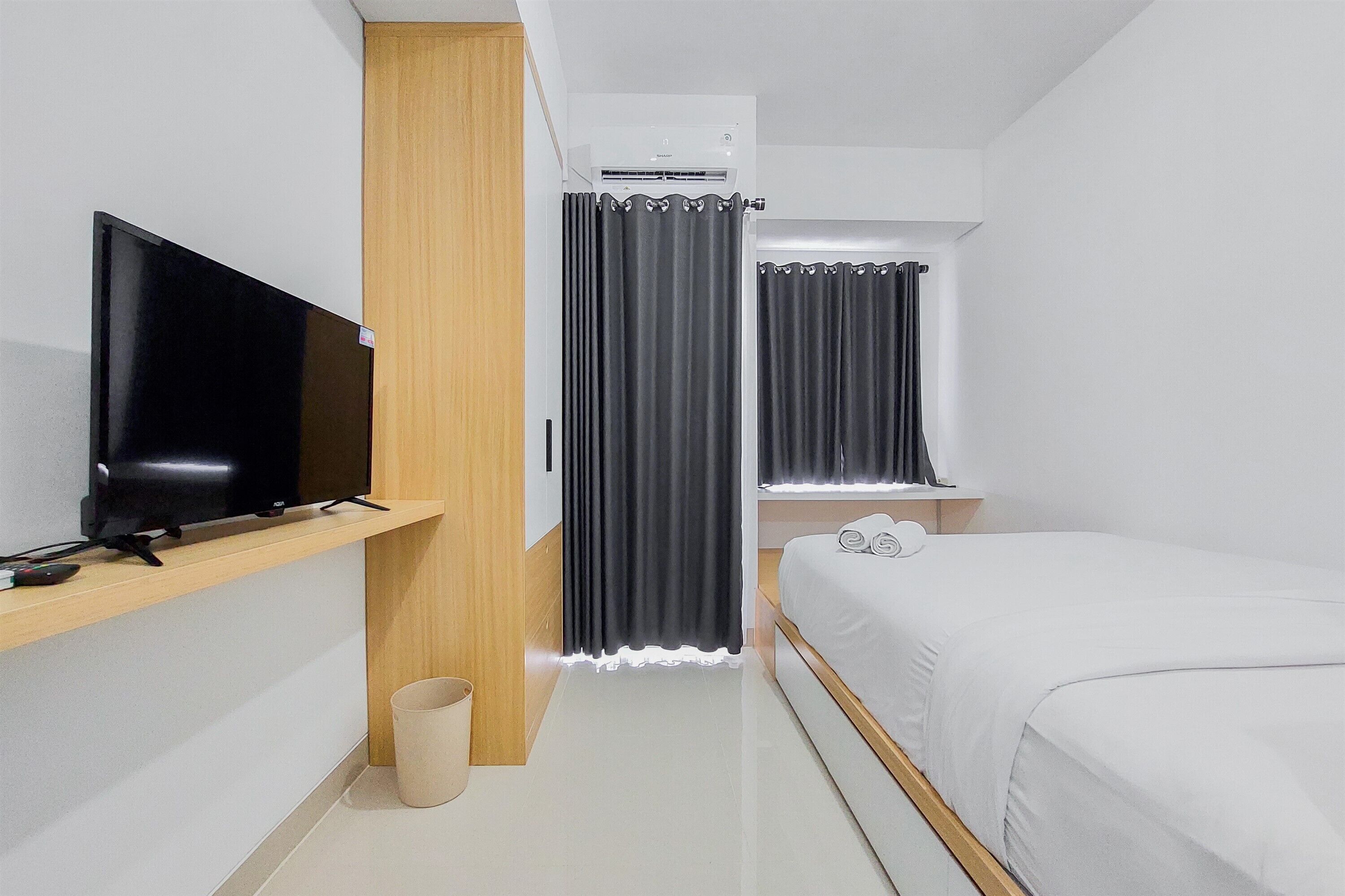 A modern hotel room with a flat-screen TV on a wooden shelf, a bed with white bedding, and a small bedside table.
