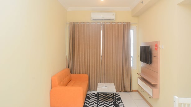 A living room with a sofa, a coffee table, a TV mounted on a wooden panel, and a window with curtains. - Elegant And Nice 2Br At Grand Palace Kemayoran Apartment (Jakarta)