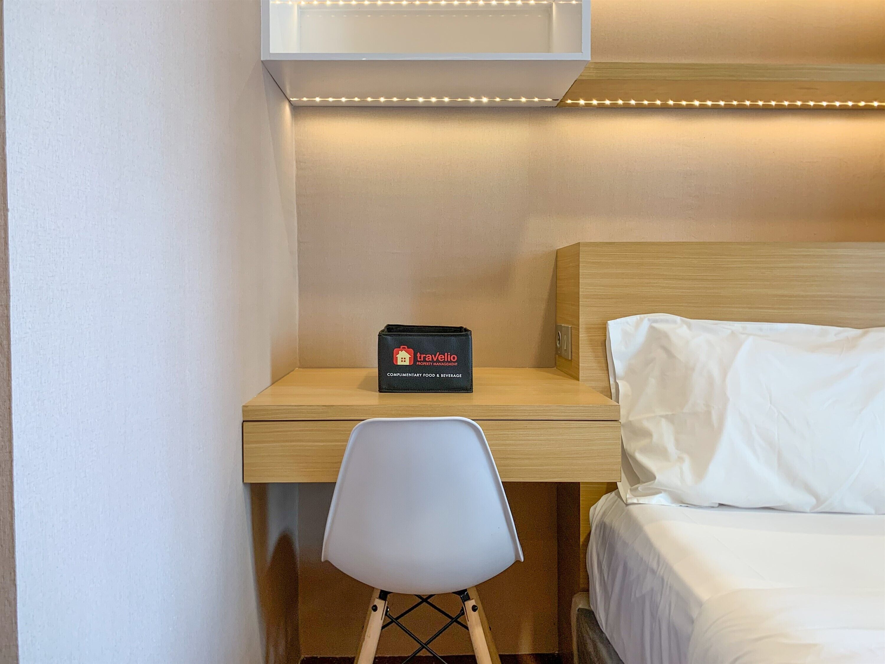 Een hotelkamer met een houten bureau, een stoel, een bed met wit beddengoed en een doosje met eten en drinken van Travelio.