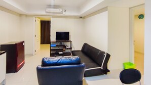 A living room with a black leather sofa, a blue chair, a television on a stand, and a small green trash bin. - New And Nice 2Br At Marquis De Lafayette Apartment (Semarang)