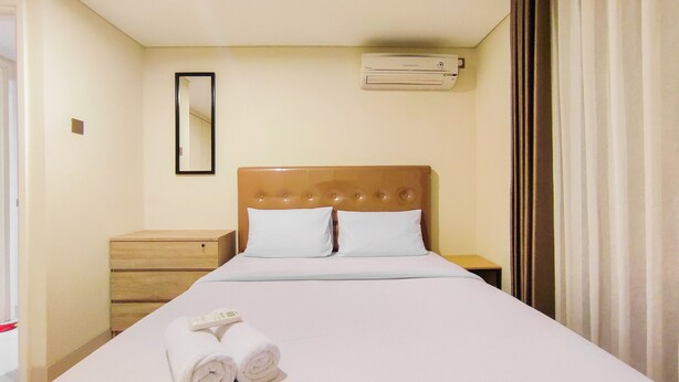 A hotel room with a large bed, a wooden nightstand, a mirror, an air conditioning unit, and a window with curtains. - New And Nice 2Br At Marquis De Lafayette Apartment (Semarang)