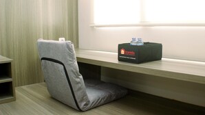 Ein modernes Hotelzimmer mit einer grau gepolsterten Bank, einem kleinen Regal mit einer Reisetasche und einem Fenster mit Jalousien.
