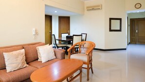 A hotel room with a sofa, a wooden chair, a dining table with chairs, an air conditioning unit, and a clock on the wall. - Extra Spacious And Homey 2Br At Kondominium Puncak Marina Apartment (Surabaya)