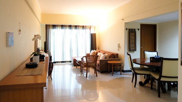 A well-lit living room with a sofa, a dining table, and chairs. There is a television on a stand, a potted plant, and a window with curtains. - Extra Spacious And Homey 2Br At Kondominium Puncak Marina Apartment (Surabaya)