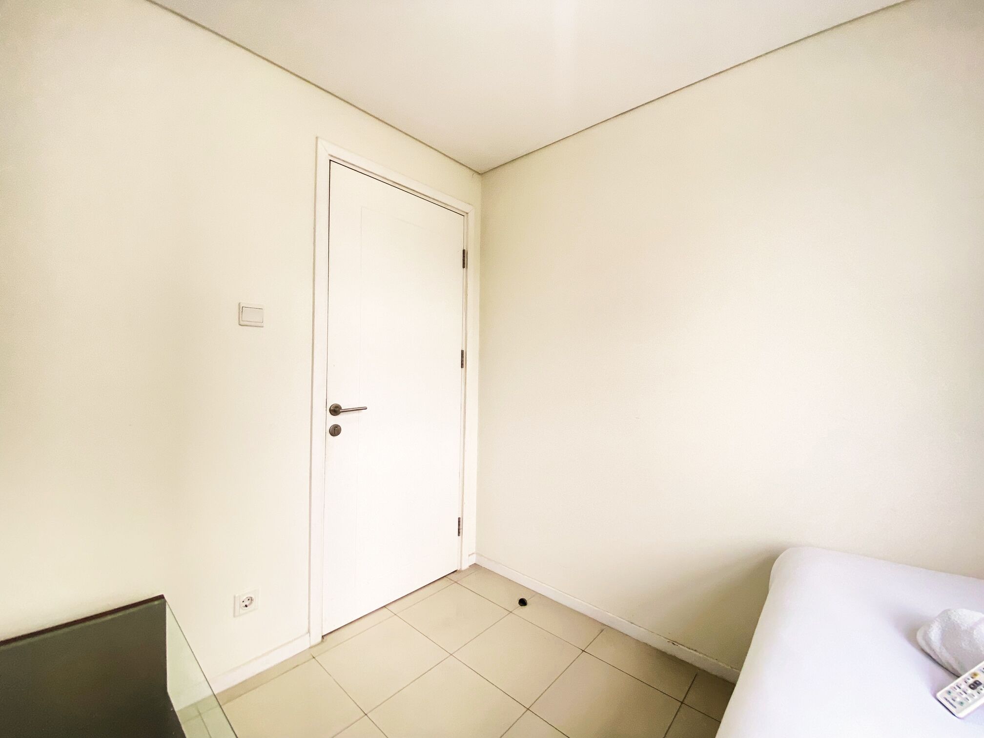 Quarto pequeno e limpo, com porta branca, uma cama e uma mesinha.