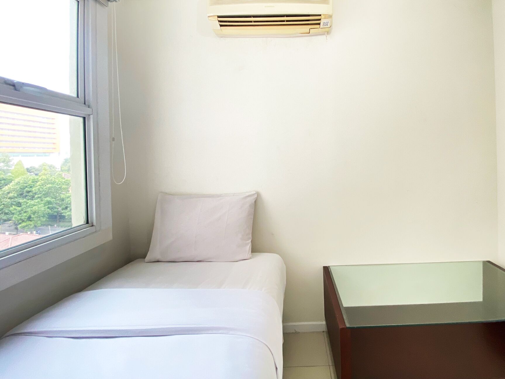 Una cama pequeña y ordenada, con un almohadón blanco y una mesita de noche con superficie de vidrio.