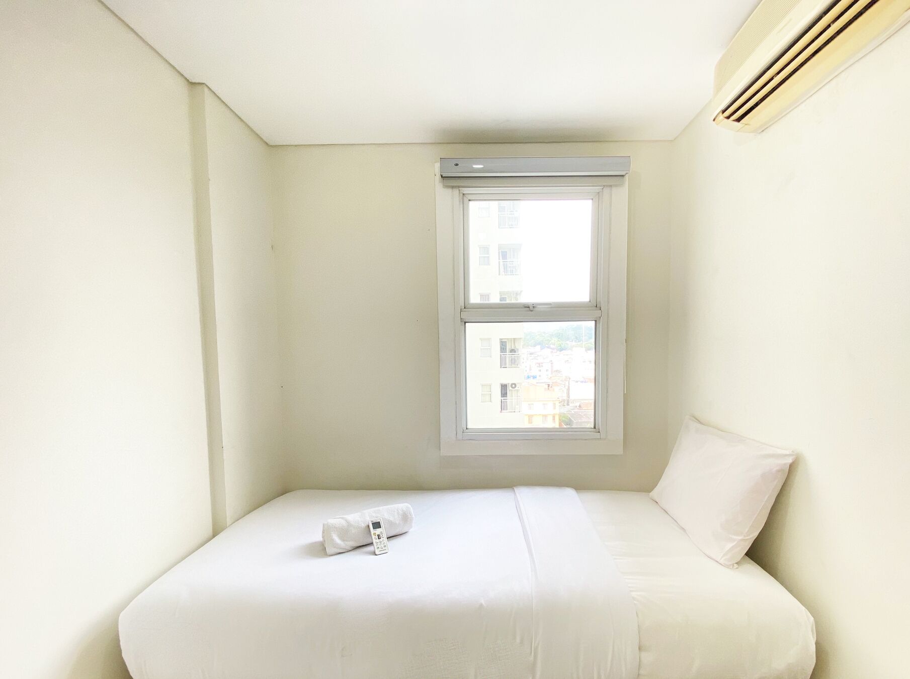 A small, white, single-bed room with a window and a wall-mounted air conditioner.