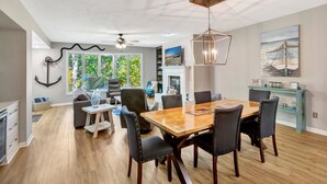 A modern dining area with a wooden table and chairs, a chandelier, and a view of greenery outside.