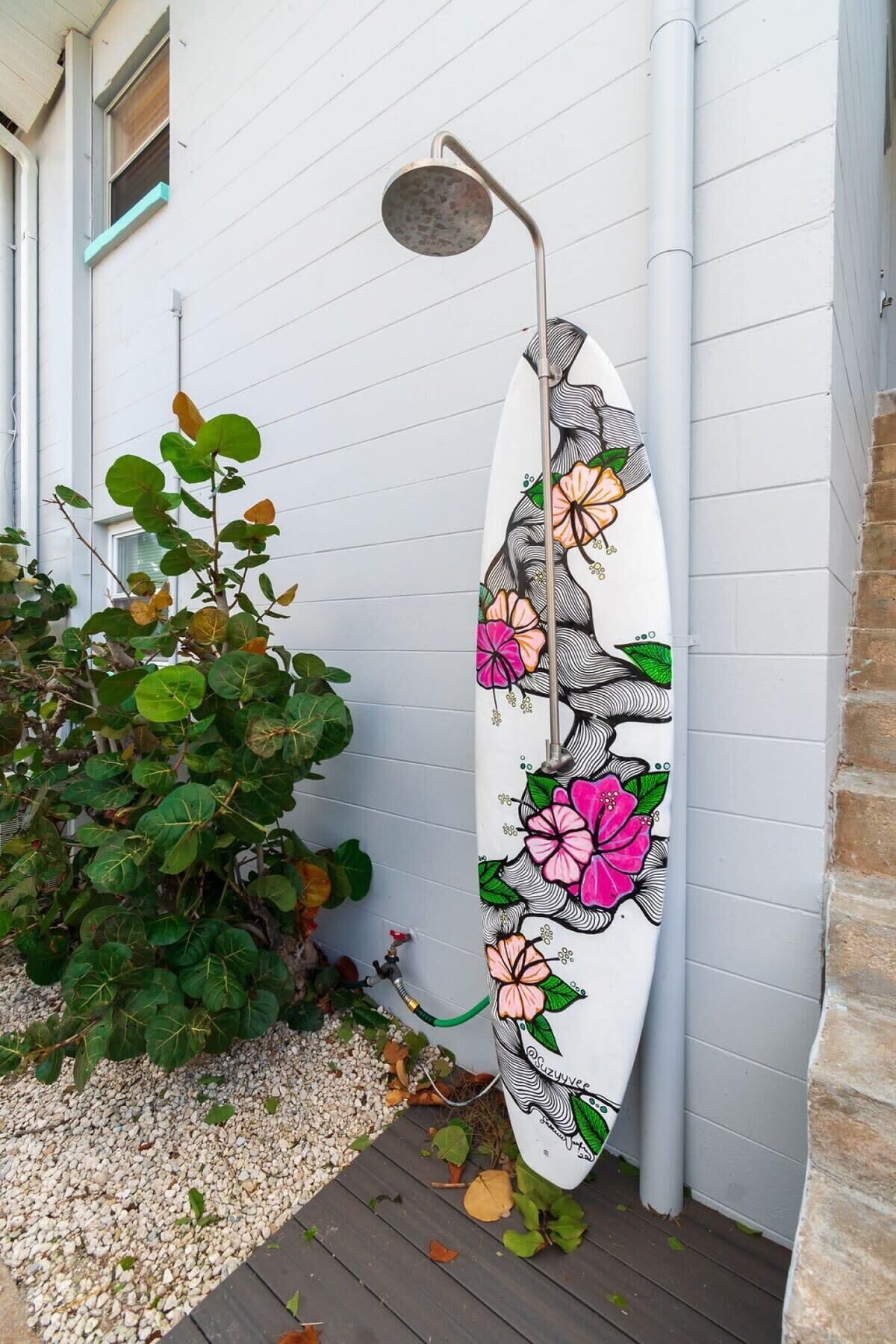 Un mur blanc orné d’une planche de surf peinte de motifs floraux, un pommeau de douche en métal et une plante placée à côté du mur.