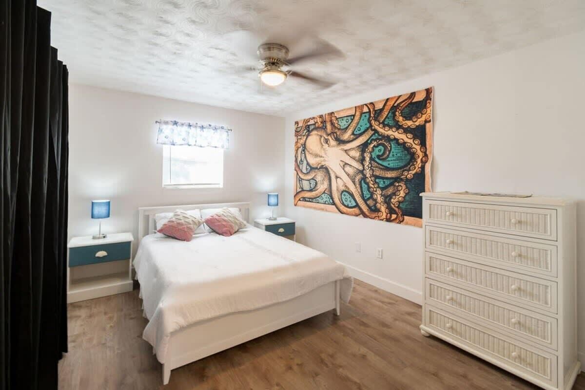 A bedroom with a bed, bedside tables, a ceiling fan, a window with curtains, and a wall painting.