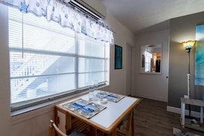 Une piĂšce avec une table, des chaises, une fenĂȘtre avec des stores, une lampe et un tableau au mur.