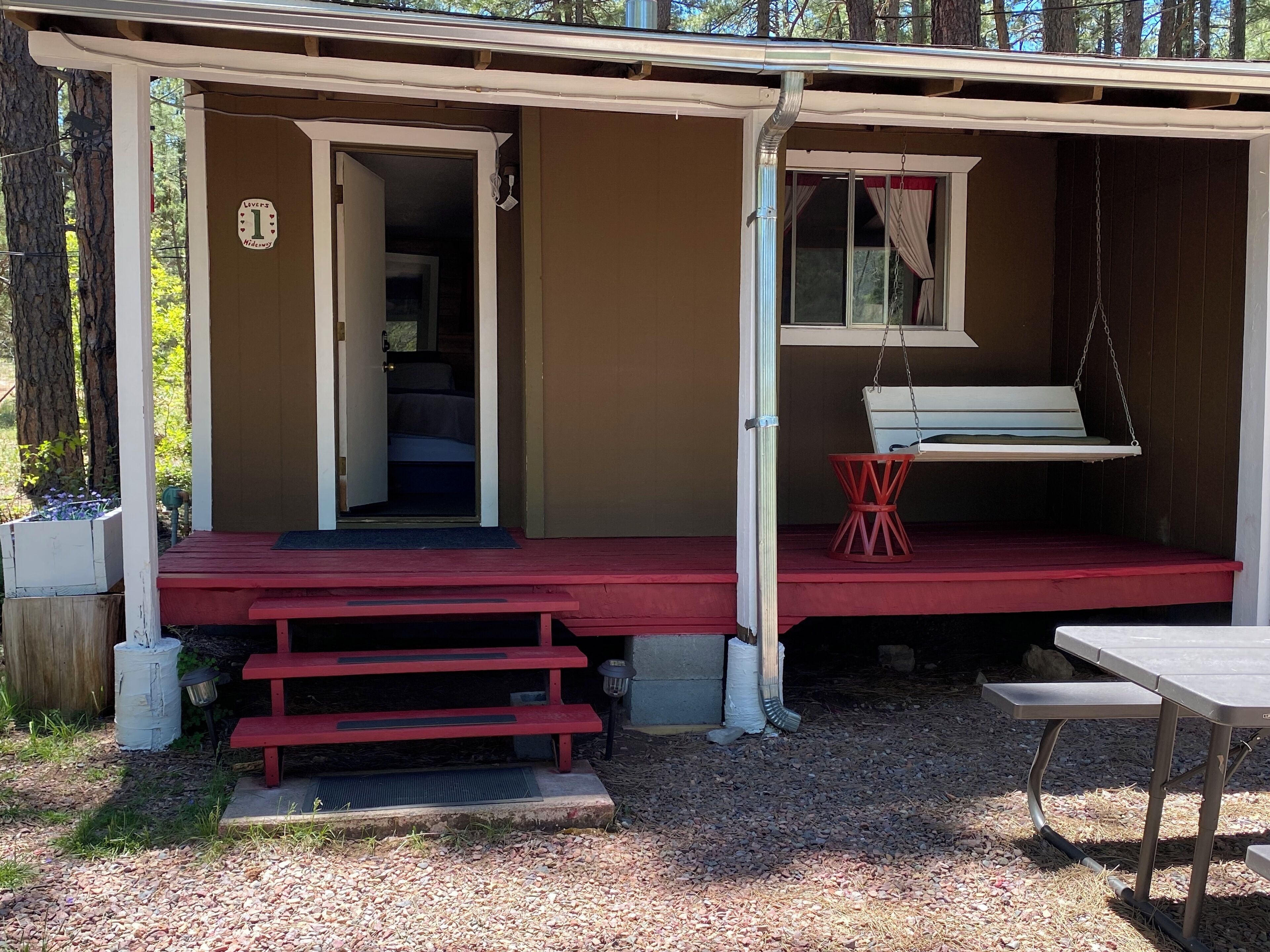 Une cabane en bois avec un porche rouge, un lit à l’intérieur, une chaise suspendue et une table.