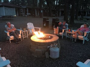 A group of people seated around a stone fire pit in an outdoor setting with wooden chairs and a portable grill.