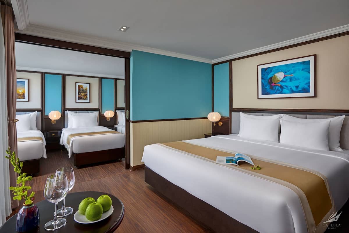 Hotelkamer met twee bedden, een grote bed, een eettafel met fruit en een glas, en een schilderij aan de muur.