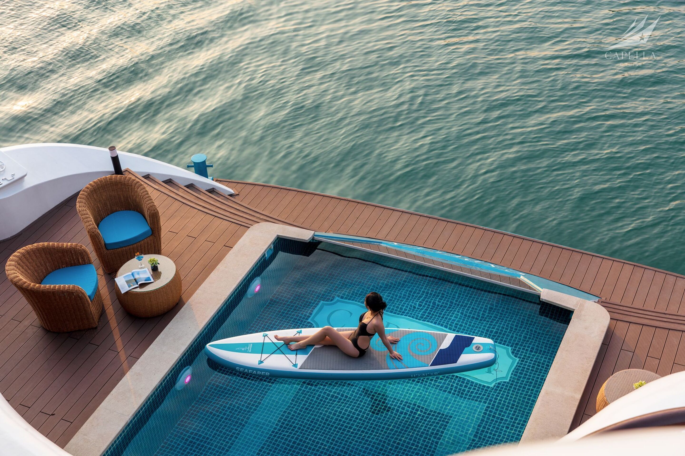 Une femme est assise sur une planche de surf dans une piscine située sur le pont d’un bateau, où se trouvent des chaises en osier et une petite table.