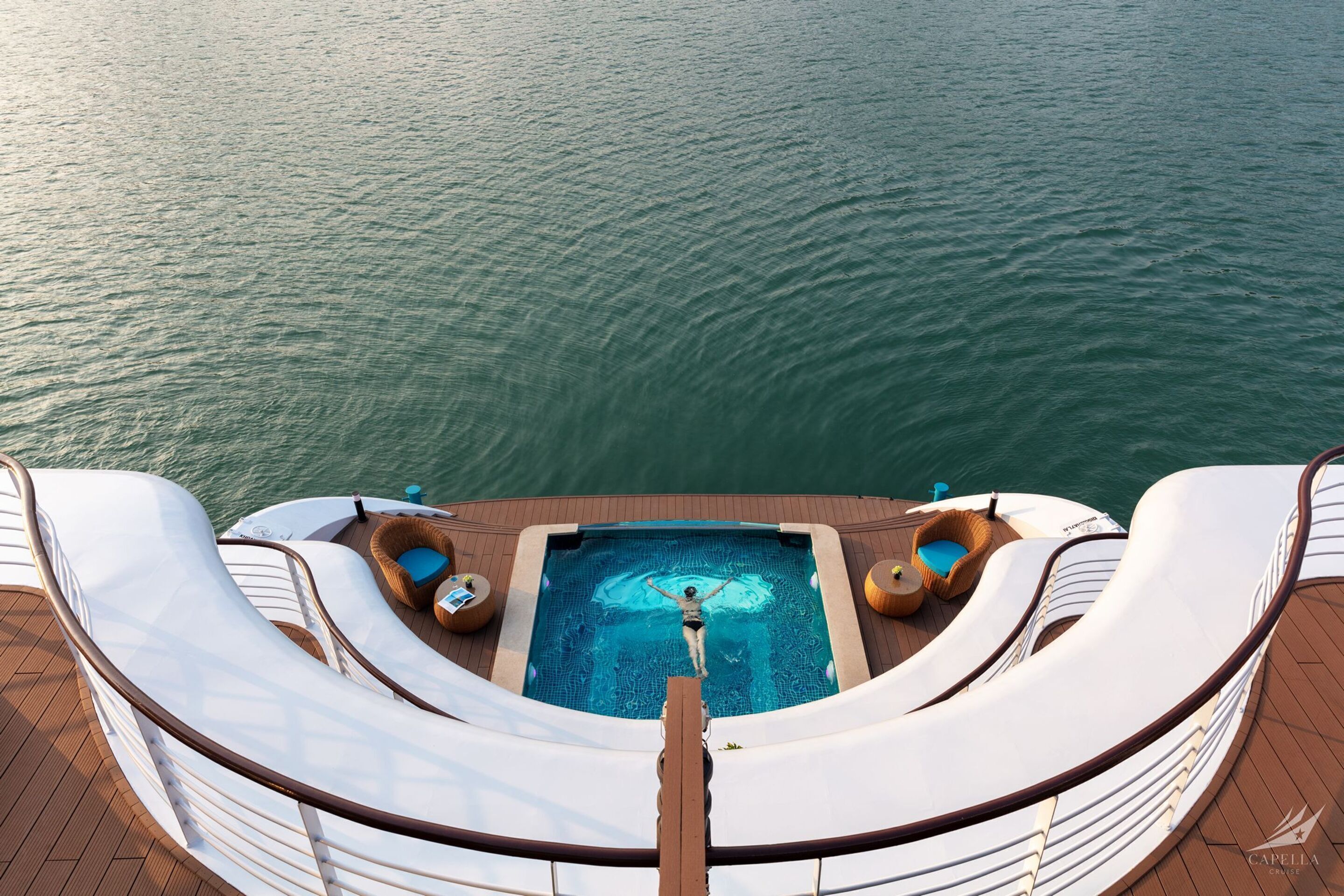 A pool area with a person swimming, surrounded by lounge chairs and a wooden deck.