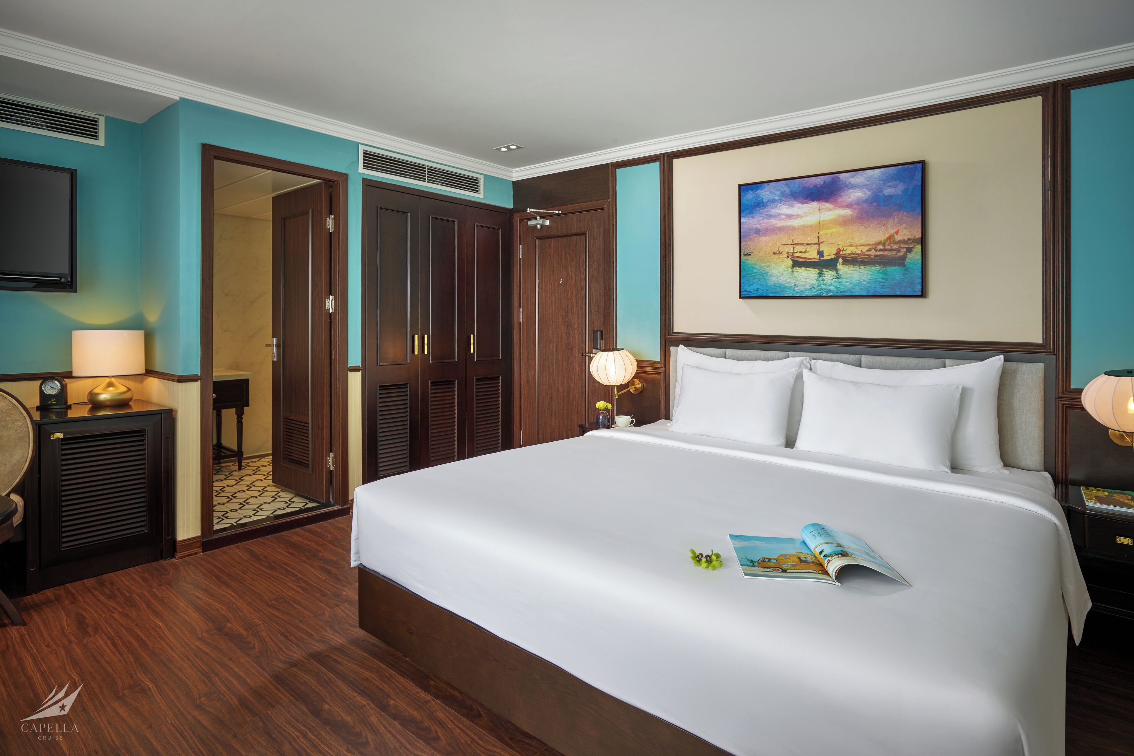 A hotel room with a large bed, a desk with a lamp, a wardrobe, and a painting on the wall.