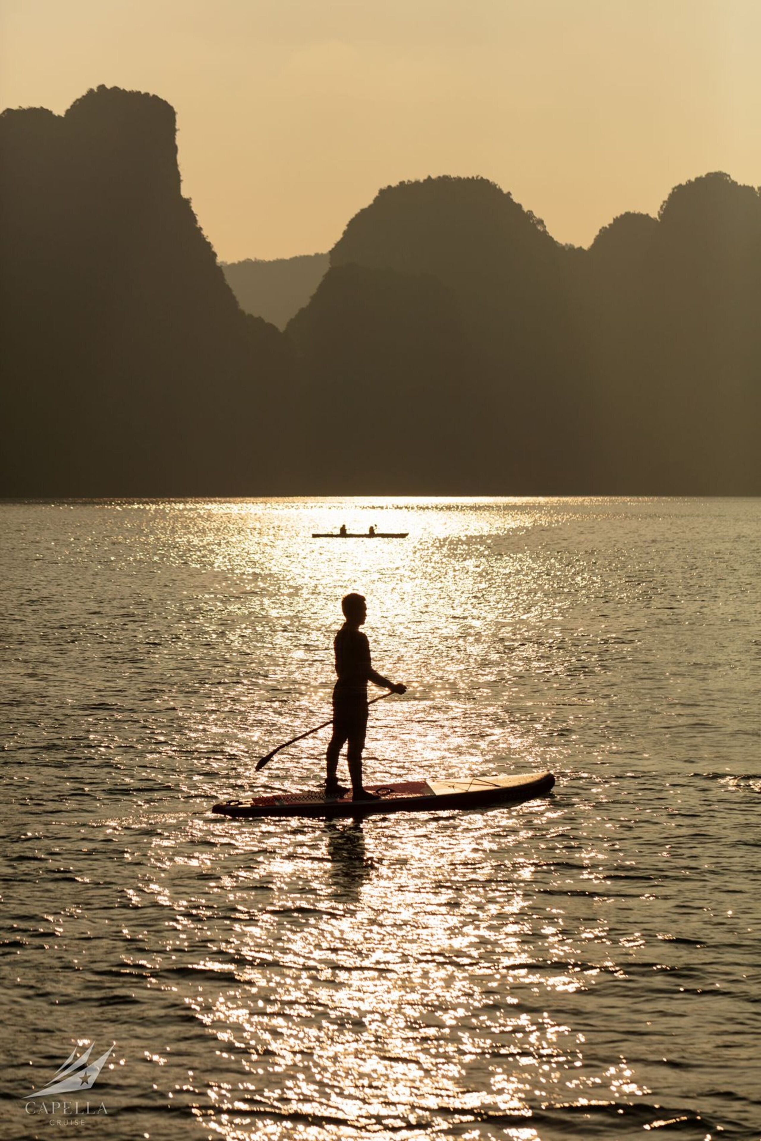 A person paddleboarding on calm water with a boat in the distance and mountains in the background.