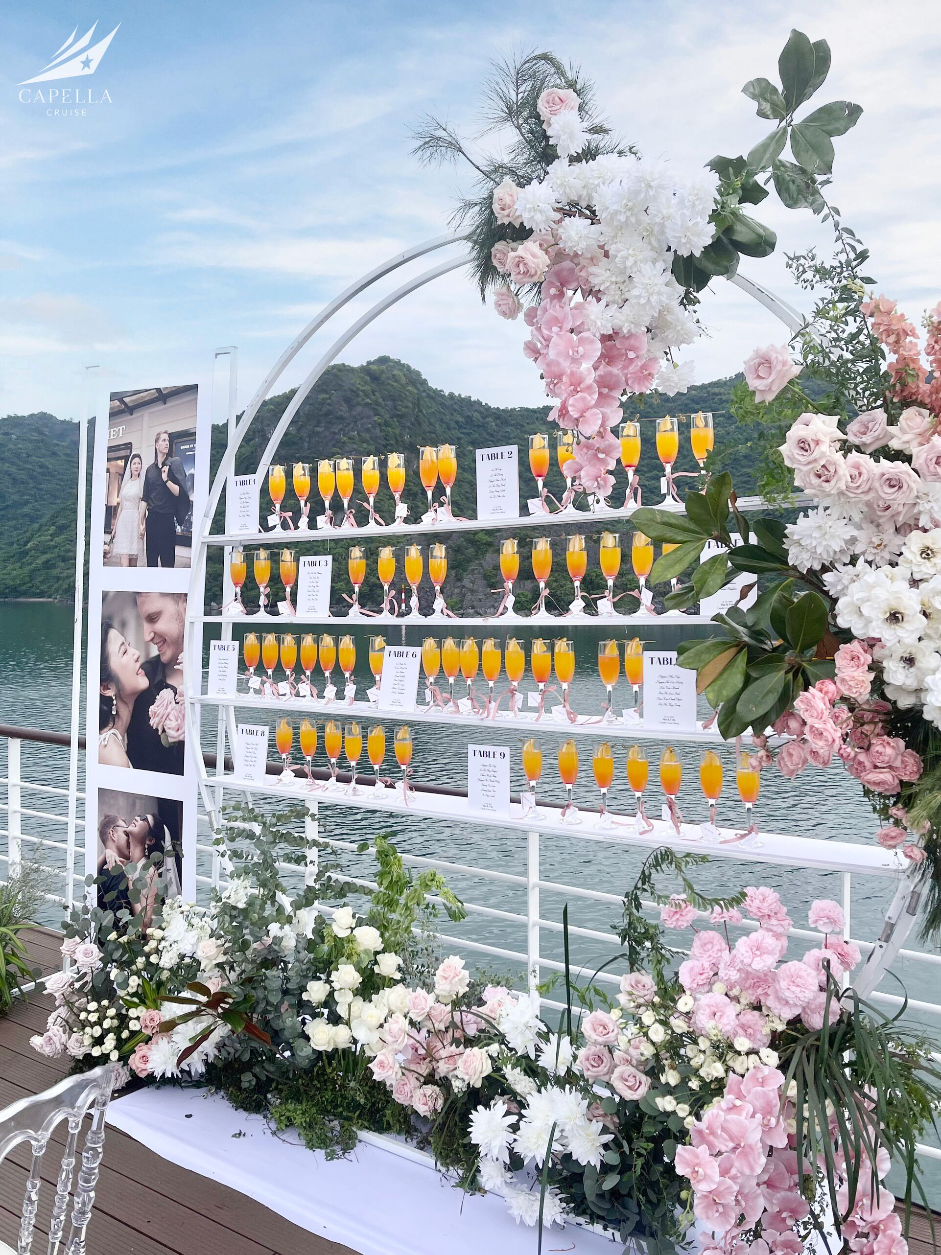 A table setup with multiple glasses of orange juice, surrounded by floral decorations and a scenic lake view.