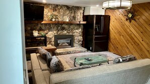 A cozy living room with a stone fireplace, wooden walls, and a modern kitchen with a built-in refrigerator. - Fully AirConditioned Resort Style home living w/Hottub,outdoor shower and  pool. (Delta)