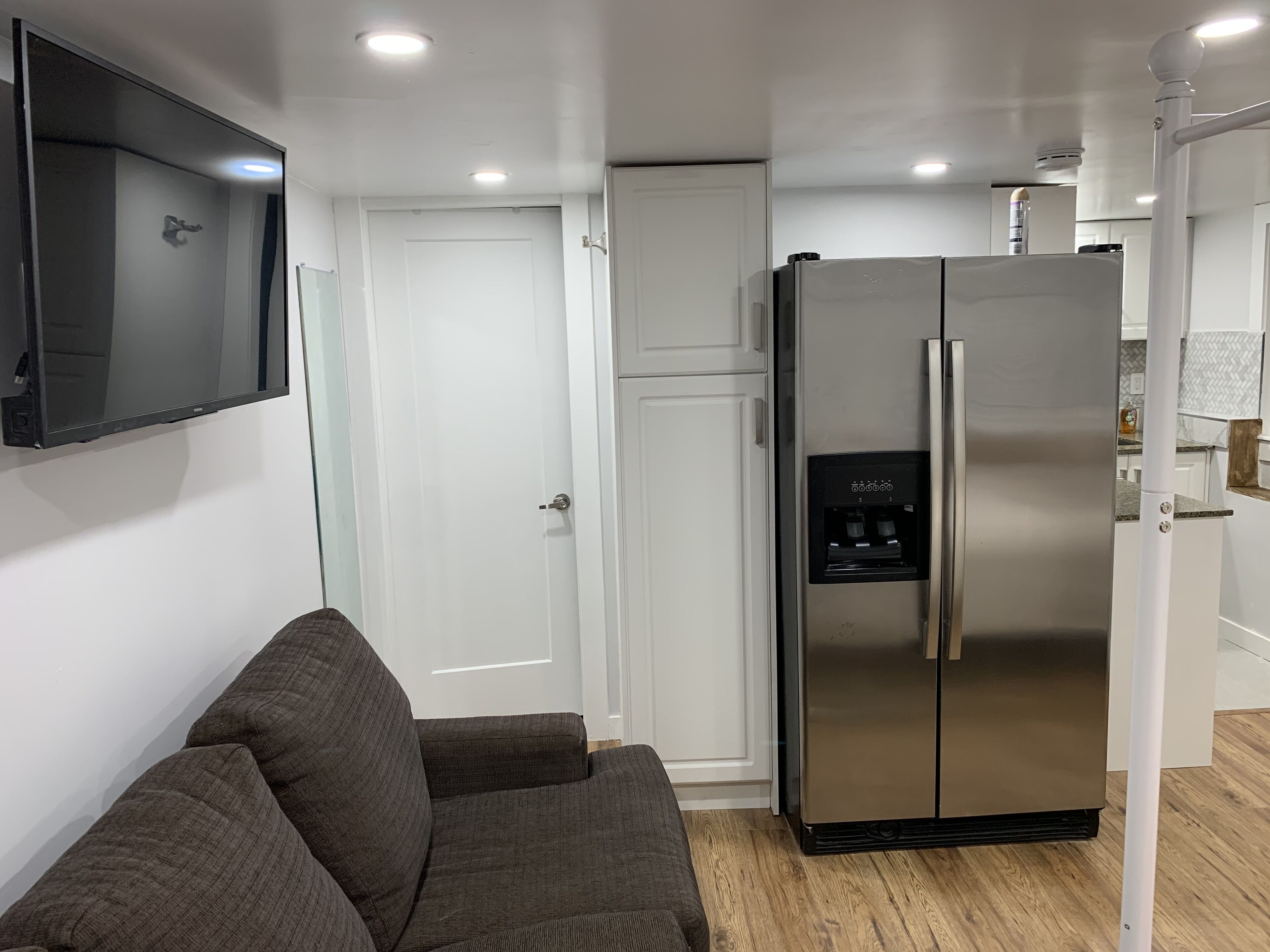 A compact kitchen with a built-in refrigerator, a flat-screen TV mounted on the wall, and a wooden floor.