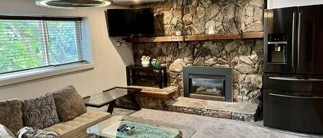 A modern living room with a stone fireplace, a flat-screen TV mounted on the wall, a black refrigerator, and a beige sofa with patterned cushions.