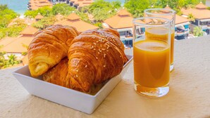Een ontbijttafel met croissants en sinaasappelsap op een balkon met uitzicht op een strand en hotelgebouwen.