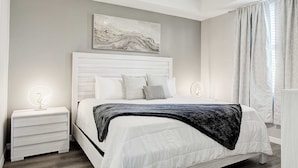 Een slaapkamer met een wit bed, een nachtkastje met een lamp, een kunstwerk aan de muur en een raam met gordijnen.