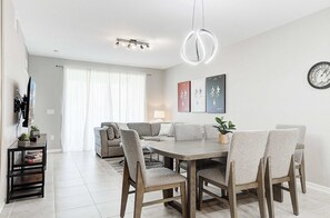 A modern dining area with a wooden table and chairs, a sofa, and a wall-mounted clock. - Resort Apartment Near Disney w/Lazy River & Gym (Kissimmee)
