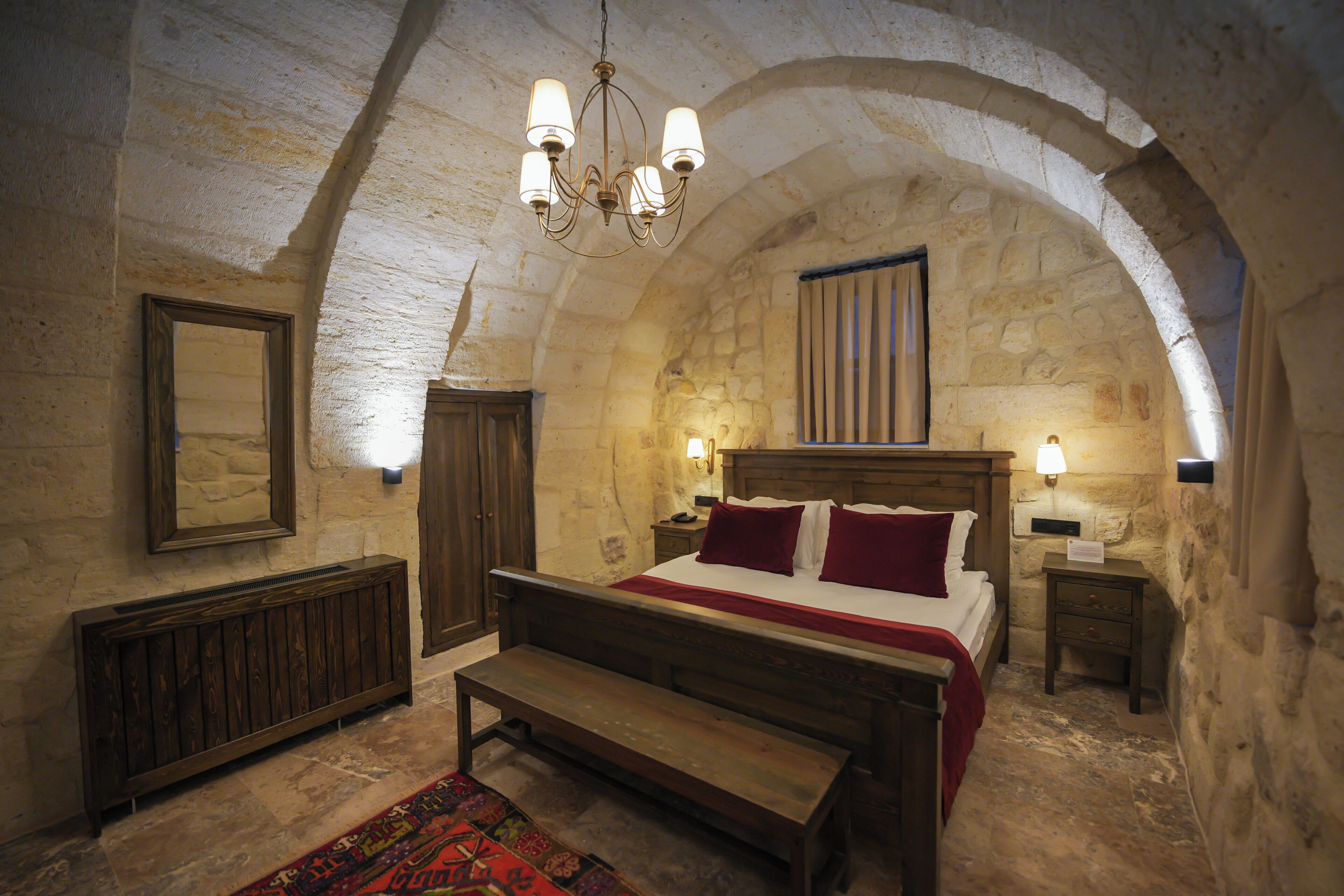 A room with a stone vaulted ceiling, a wooden bed with red bedding, a bench, a nightstand, and a wall-mounted lamp.