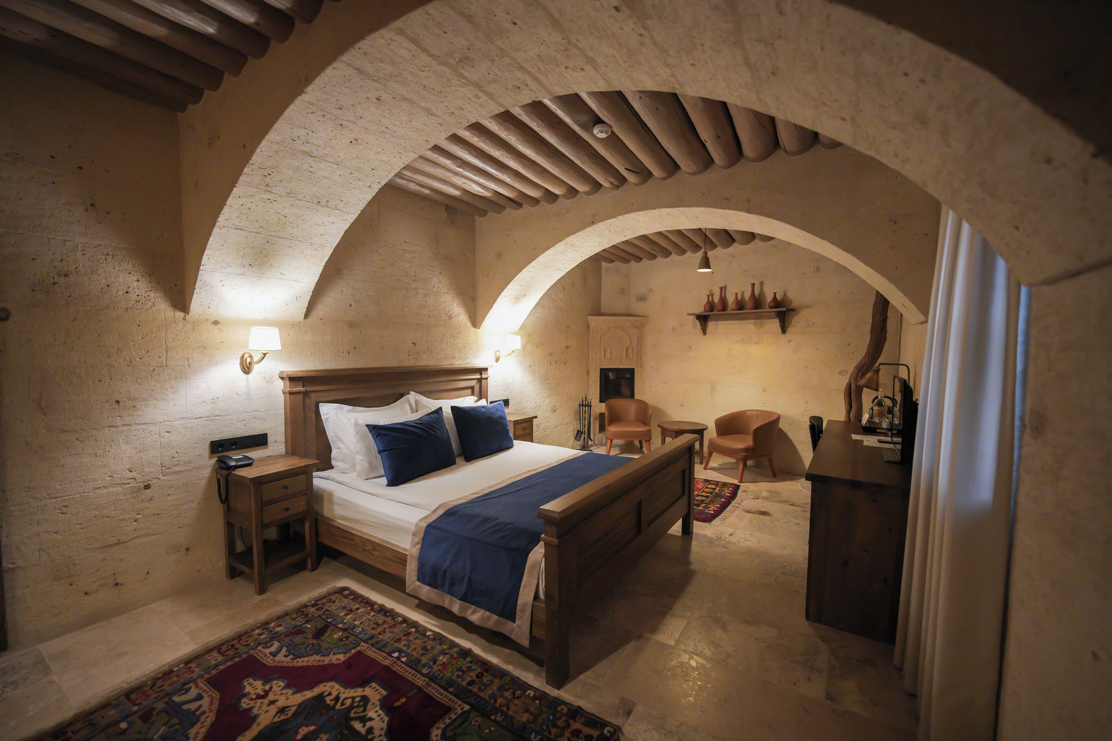 A bedroom with a wooden bed, a nightstand, a chair, a desk, and a small shelf with decorative items.