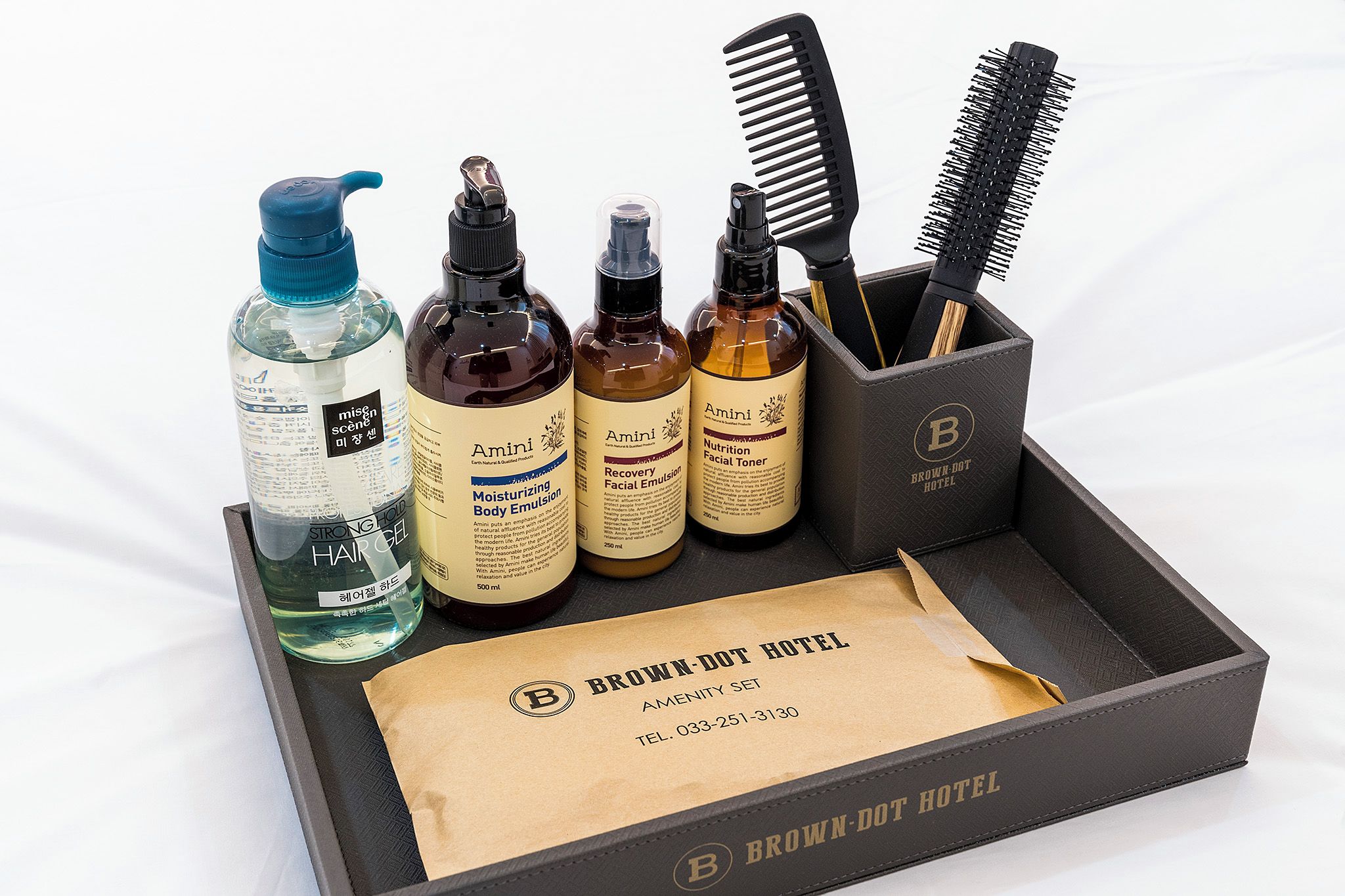 Un conjunto de artículos de aseo que incluye un bote de gel para el cabello, tónico facial y un peine, presentado en un set de amenidades de hotel.