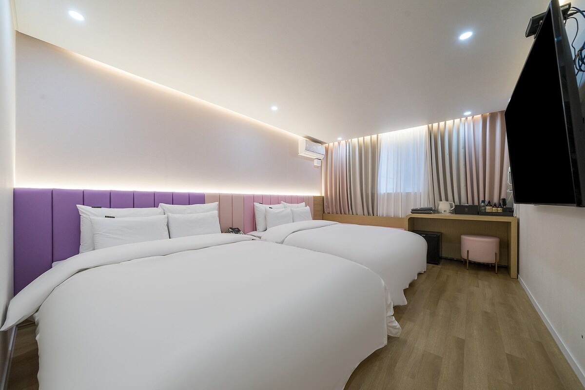 A hotel room with two beds, a desk, a chair, a TV, and a window with curtains.