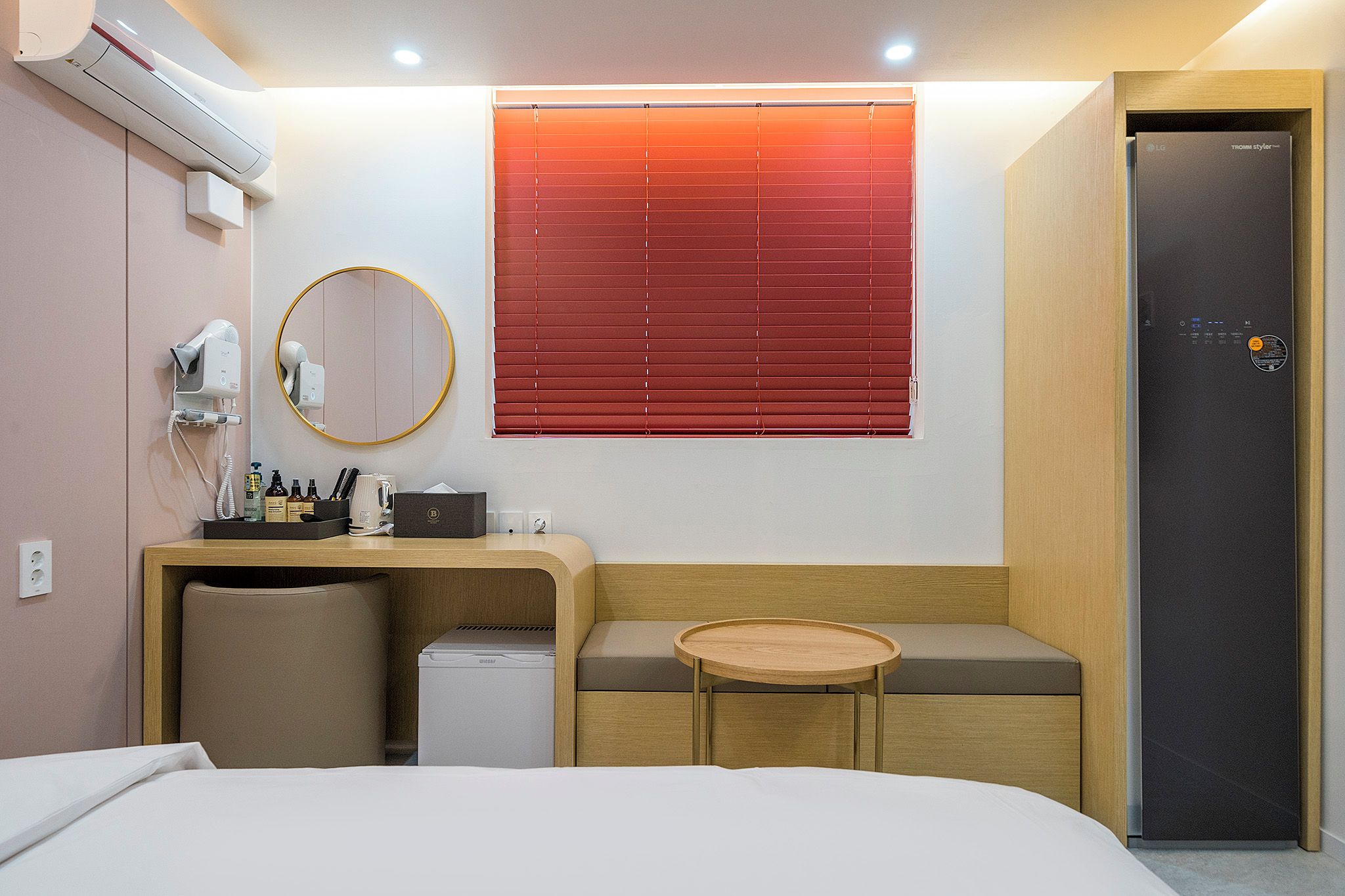 A hotel room with a bed, a desk with a chair, a small table, a mirror, a hairdryer, and a refrigerator.