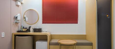 A hotel room with a bed, a desk with a chair, a small table, a mirror, a hairdryer, and a refrigerator.