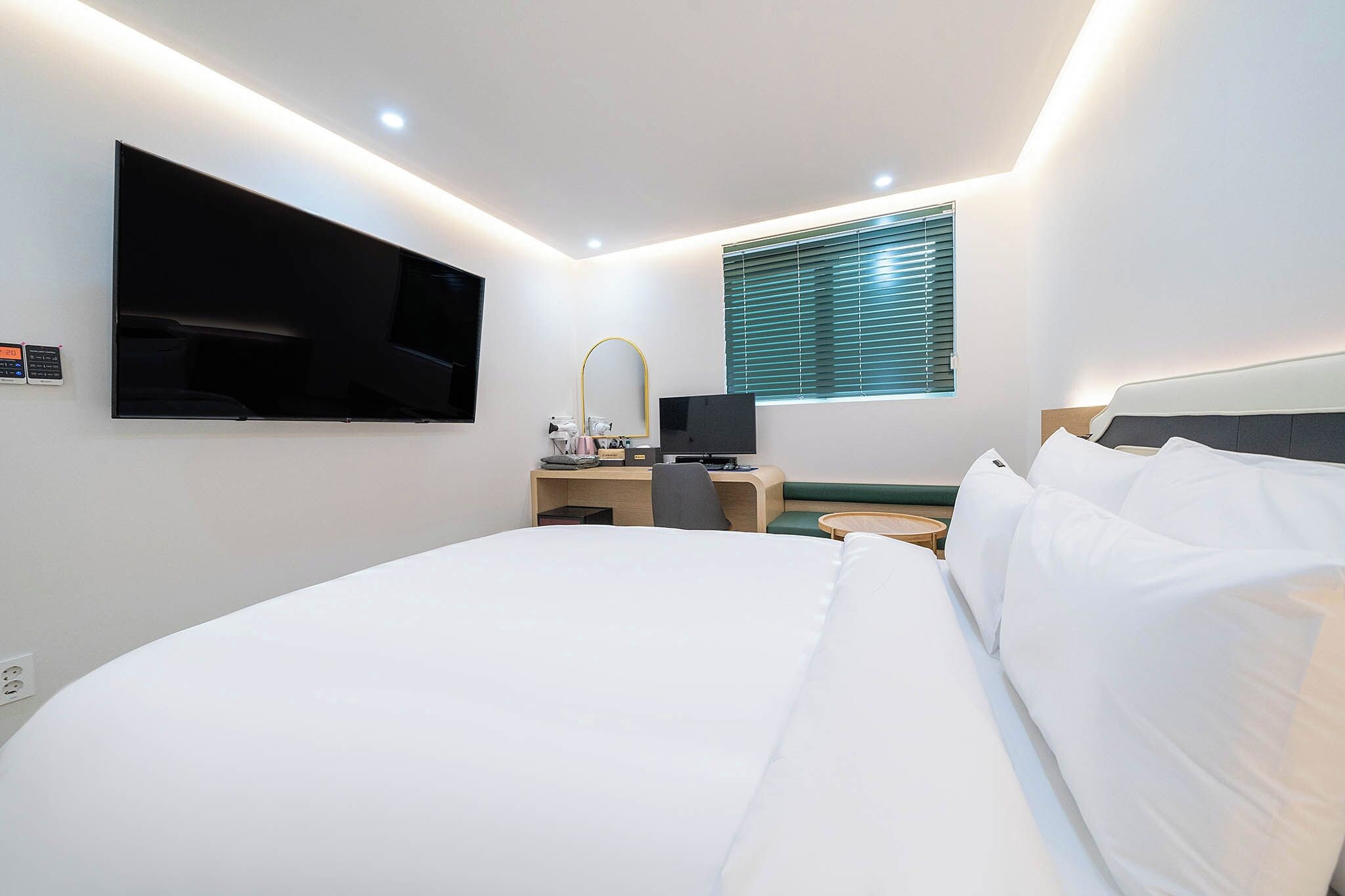 Een hotelkamer met een groot bed, een bureau met een computer, een aan de muur bevestigde televisie en een raam met jaloezieën.