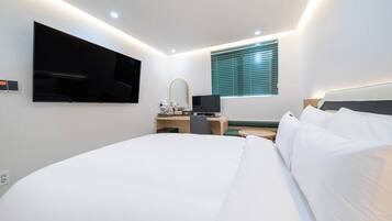 Una habitación de hotel con una cama grande, un escritorio con computadora, un televisor montado en la pared y una ventana con persianas.
