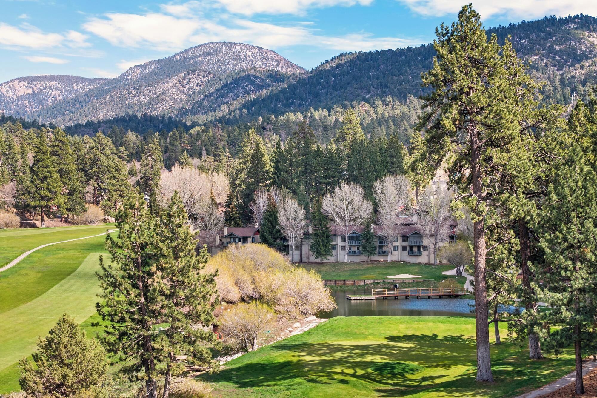 A golf course with a lake, a wooden bridge, and a building with a mountain backdrop.