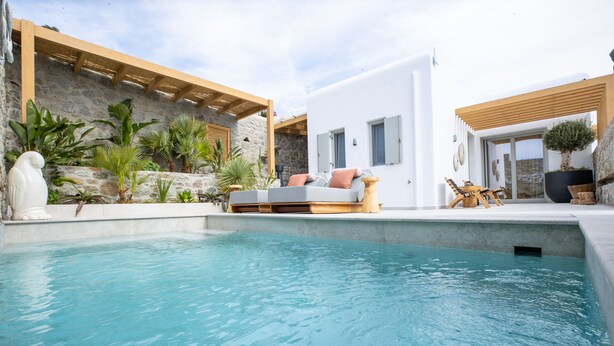 A modern outdoor pool area with a stone wall, wooden pergola, and a white building with a grey sofa and cushions. - Brilliance of Mykonos (Mykonos)