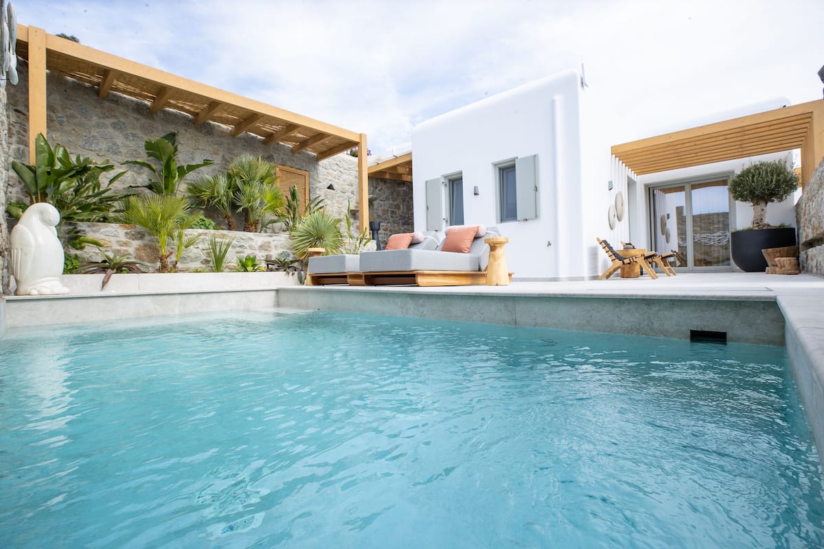 A modern outdoor pool area with a stone wall, wooden pergola, and a white building with a grey sofa and cushions.