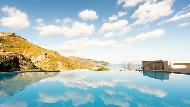 Una piscina infinita de color azul intenso con vista a un paisaje montañoso y un edificio a lo lejos.