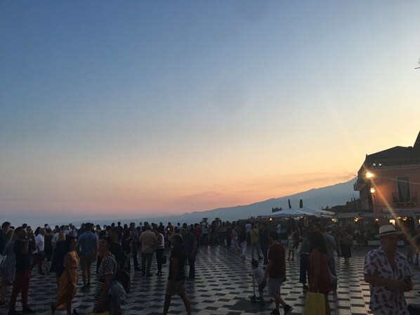 A crowded square with a checkered pavement, a mountain in the background, and a building with a balcony and lights.