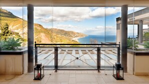 Un balcón con barandilla de vidrio, que ofrece una vista panorámica de un paisaje costero con colinas y el mar.