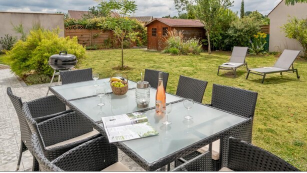 Een glazen tafel met rieten stoelen, een barbecue en een klein houten bouwwerk in een achtertuin.